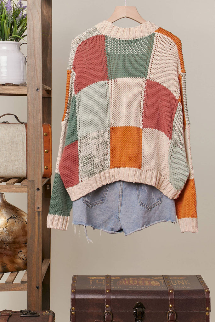 Colorful patchwork sweater on a hanger with a wooden shelf and suitcase in the background