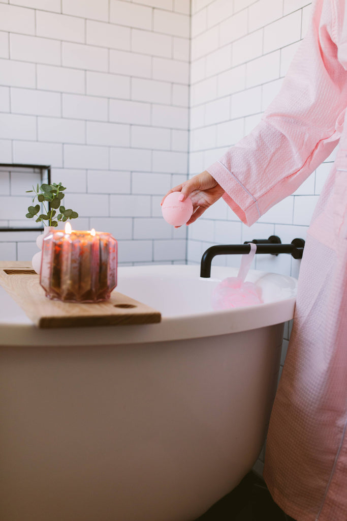 Person in a pink robe by a bathtub with a candle and plant, in a bathroom setting.