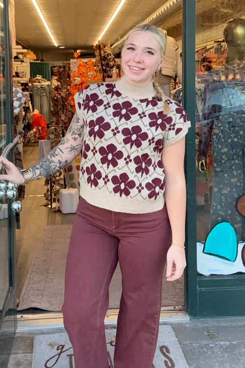 Woman wearing a floral sweater and maroon pants standing in front of a store entrance.