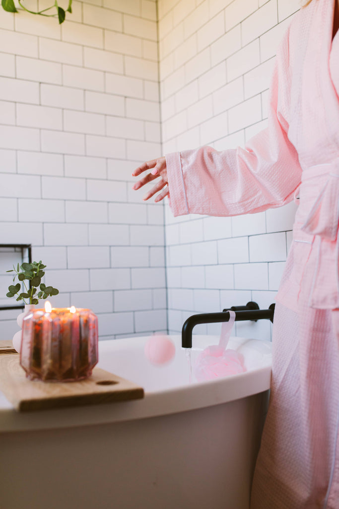 Person in a pink robe by a bathtub with candles and a plant in a bathroom.