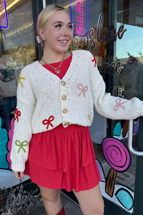Woman wearing a white cardigan with colorful bows and a red skirt in front of a colorful store entrance.