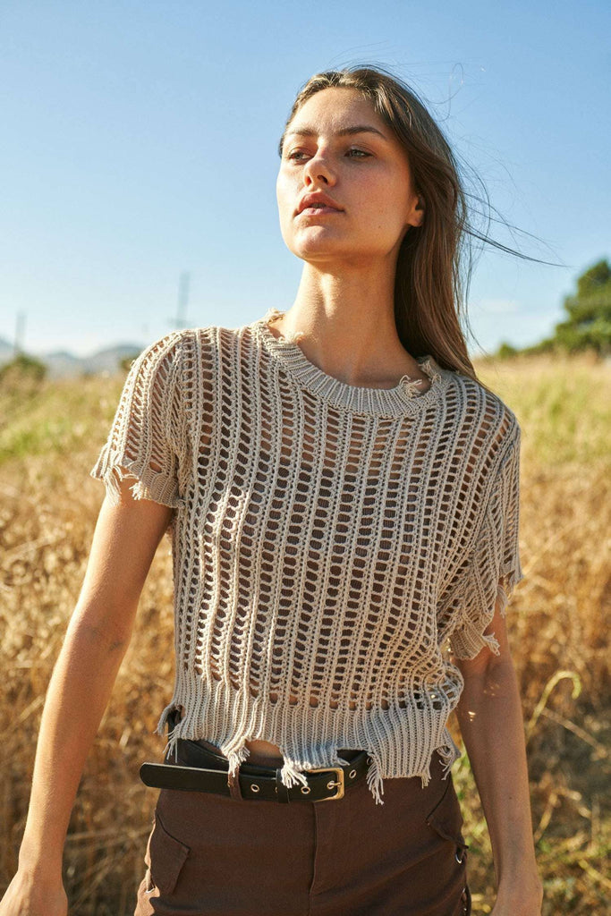 Woman wearing a beige crochet top in a field with a clear blue sky.