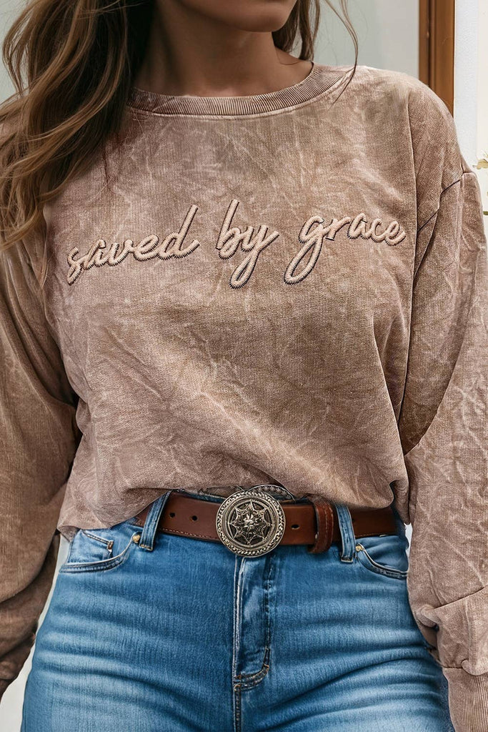 Person wearing a brown sweatshirt with 'saved by grace' text, blue jeans, and a brown belt with a large buckle.