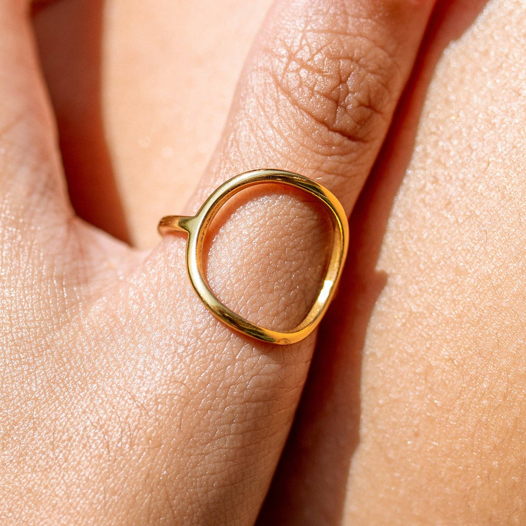 A close-up of a hand wearing a minimalist open-circle gold ring on the middle finger. The ring has a smooth, organic shape and a polished 18K gold finish, resting against sunlit, naturally textured skin.