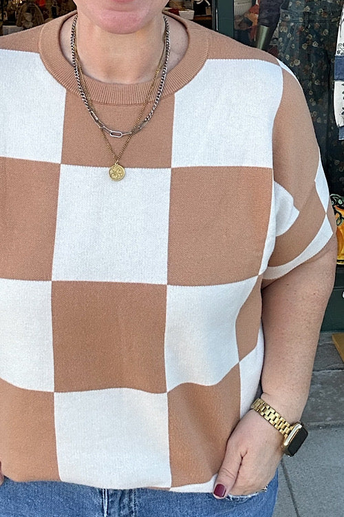Person wearing a checkered brown and white shirt with layered necklaces and a watch.