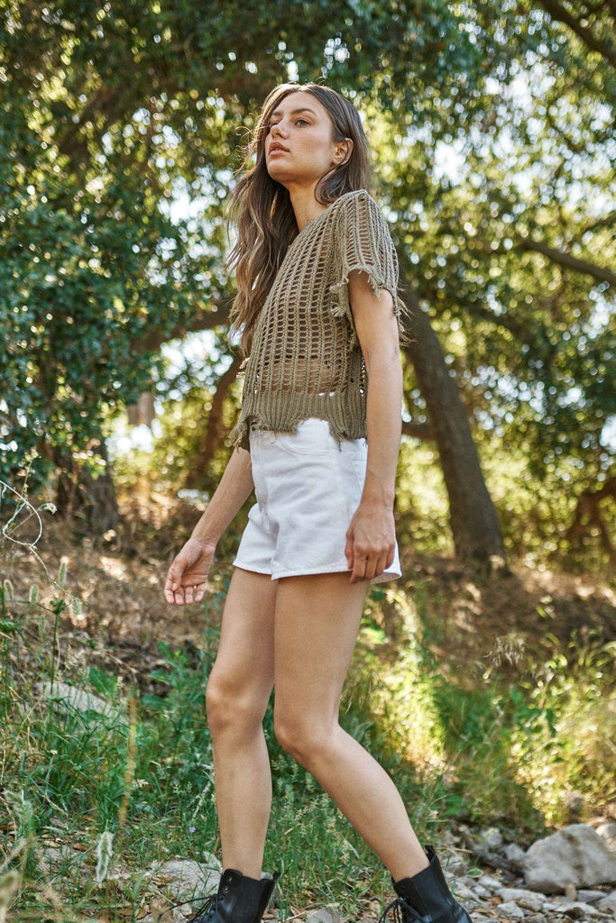Woman in a green crochet top and white shorts standing in a natural setting with trees and greenery.
