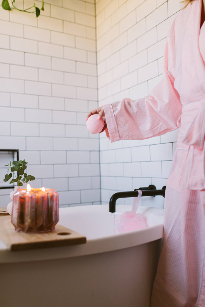 Person in a pink robe standing by a bathtub with candles and a plant in a tiled bathroom.