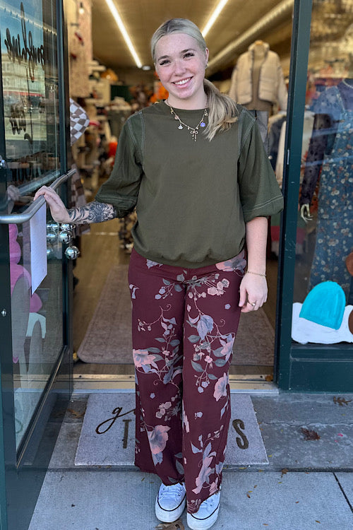 Person wearing an olive green top and maroon floral pants standing outside a store.