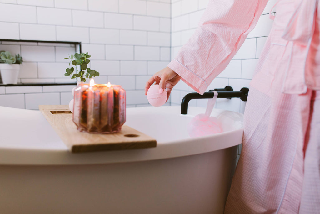 Person in a pink robe by a bathtub with a candle and plant