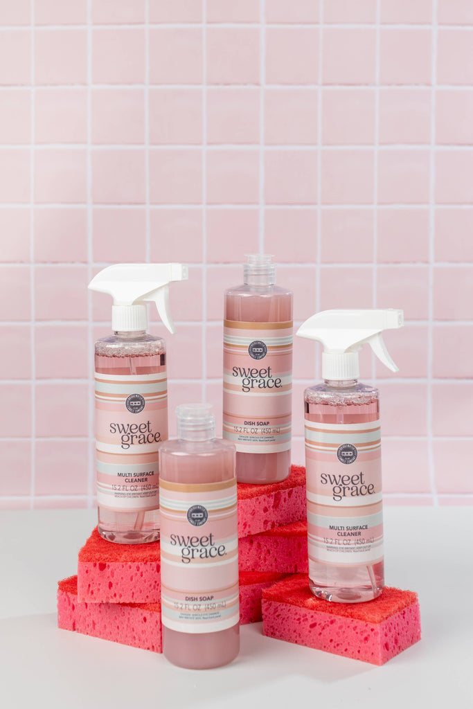 Three bottles of 'sweet grace' cleaning products on red sponges against a pink tiled wall.