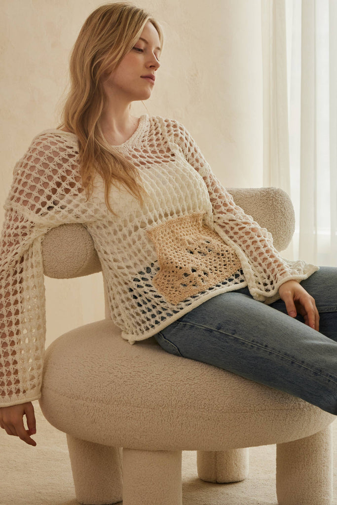 Woman sitting on a beige chair wearing a beige crochet sweater and blue jeans.