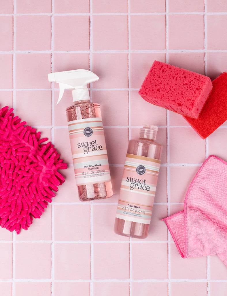 Two bottles of 'sweet grace' cleaning products on a pink tiled surface with cleaning tools.