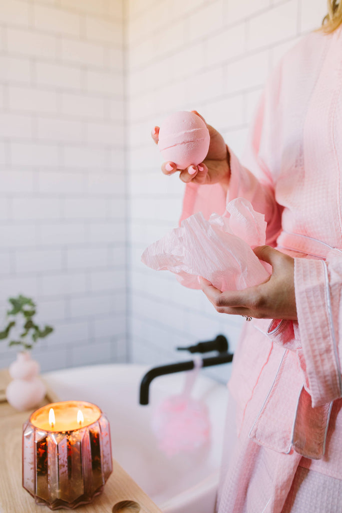 Person holding pink bath bombs in a bathroom setting with a candle and bathtub.