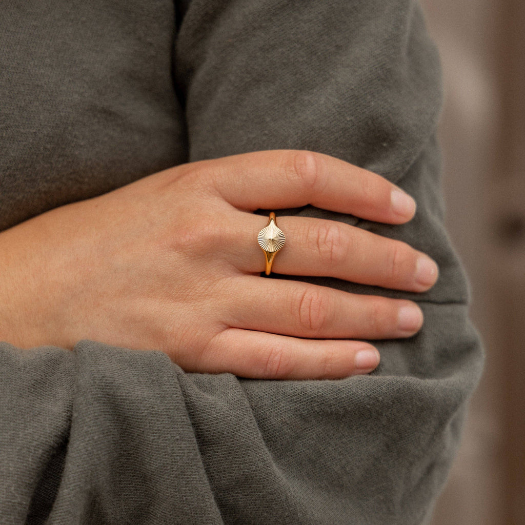 Hand wearing a gold ring with a textured design, against a neutral background