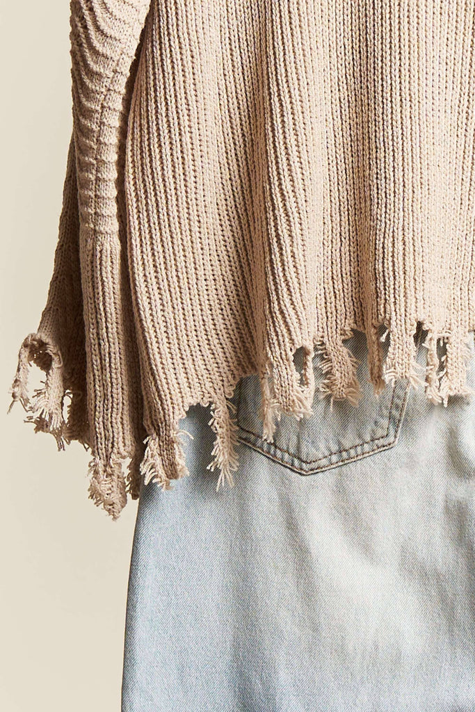 Close-up of a beige knitted sweater with frayed edges over light blue jeans on a neutral background