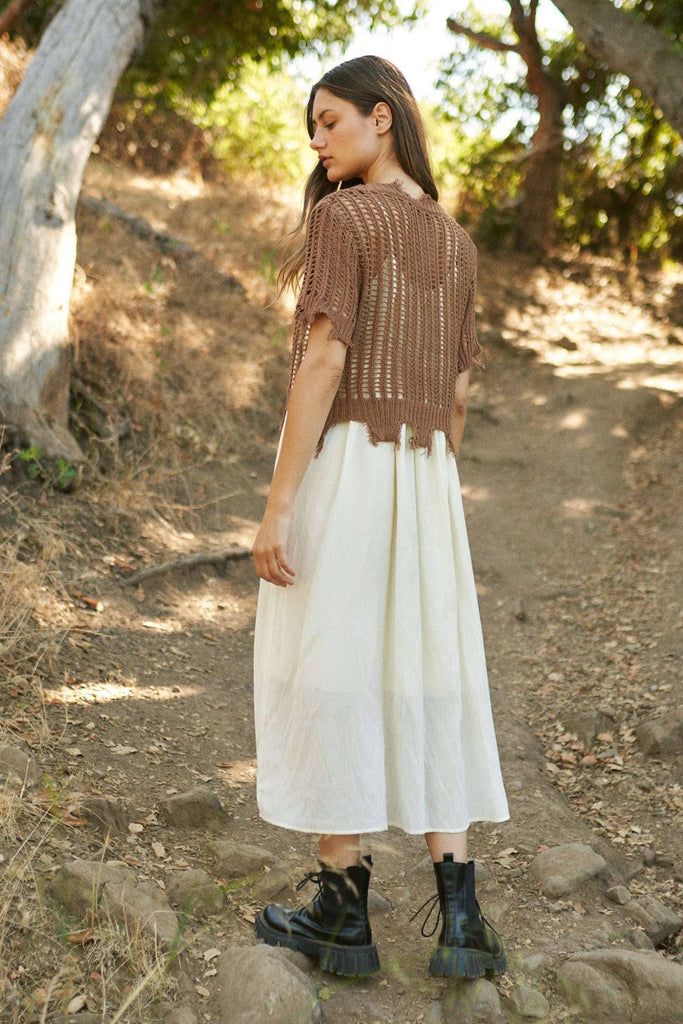 Woman in a brown knit top and white skirt standing on a dirt path with trees in the background