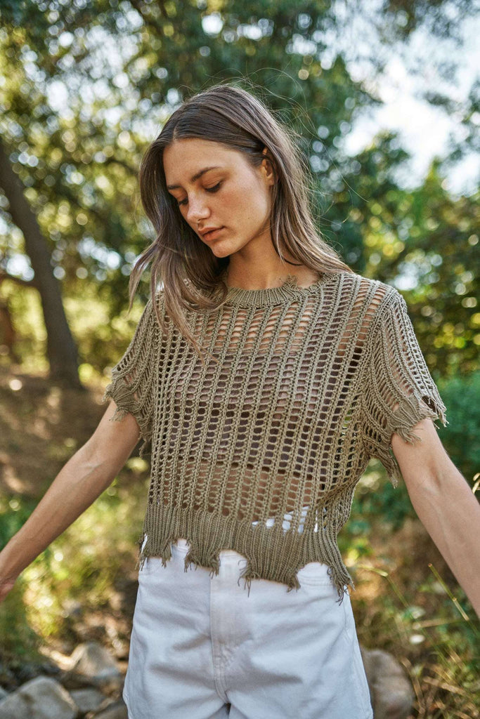 Woman wearing a green knitted top and white pants standing outdoors with trees in the background
