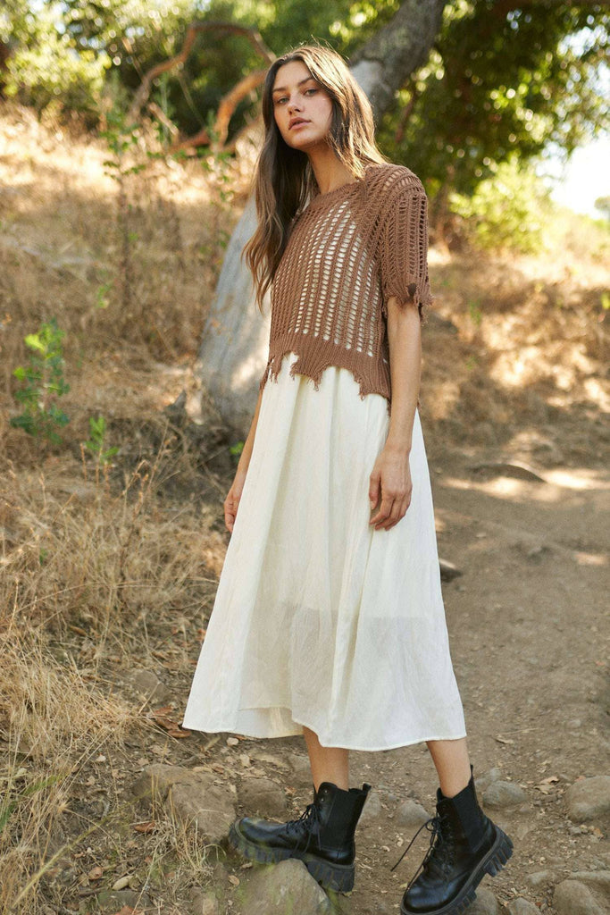Woman wearing a brown crochet top and white dress standing in a natural setting.