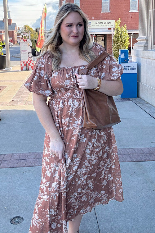 Woman in a floral dress holding a brown bag on a city street.