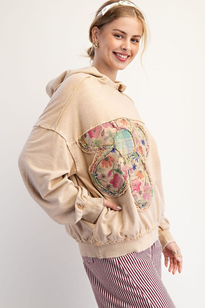 A latte-colored washed terry pullover hoodie with a large flower patch on the front made of mixed floral and plaid fabrics, and exposed stitched seams across the chest.