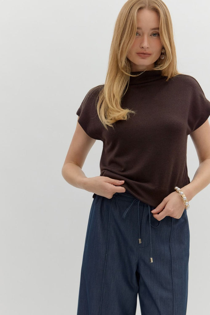 A woman with long blonde hair wears a solid brown, short-sleeve, mock neck knit top, styled with dark blue pants.