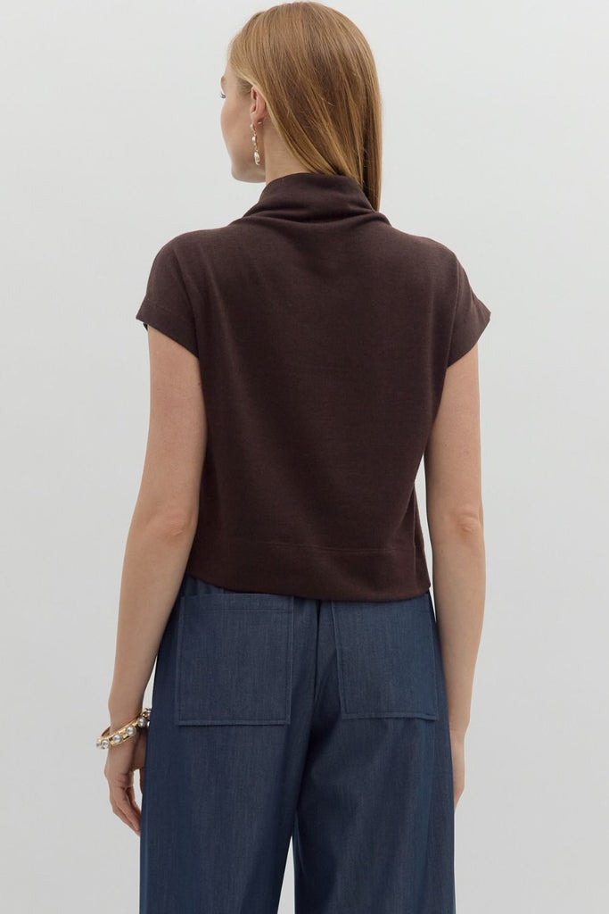 A woman with long blonde hair wears a solid brown, short-sleeve, mock neck knit top, styled with dark blue pants.