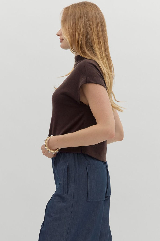 A woman with long blonde hair wears a solid brown, short-sleeve, mock neck knit top, styled with dark blue pants.