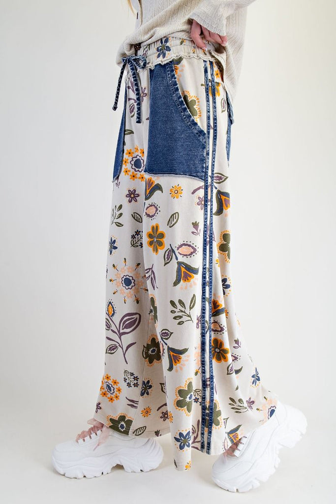 Annelise floral patchwork wide-leg pants on an ecru background with denim pocket accents, styled with a drawstring waist and chunky white sneakers.