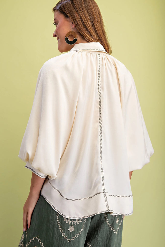 A cream satin button-down blouse with three-quarter balloon sleeves and black zigzag contrast stitching on the front placket and hem.
