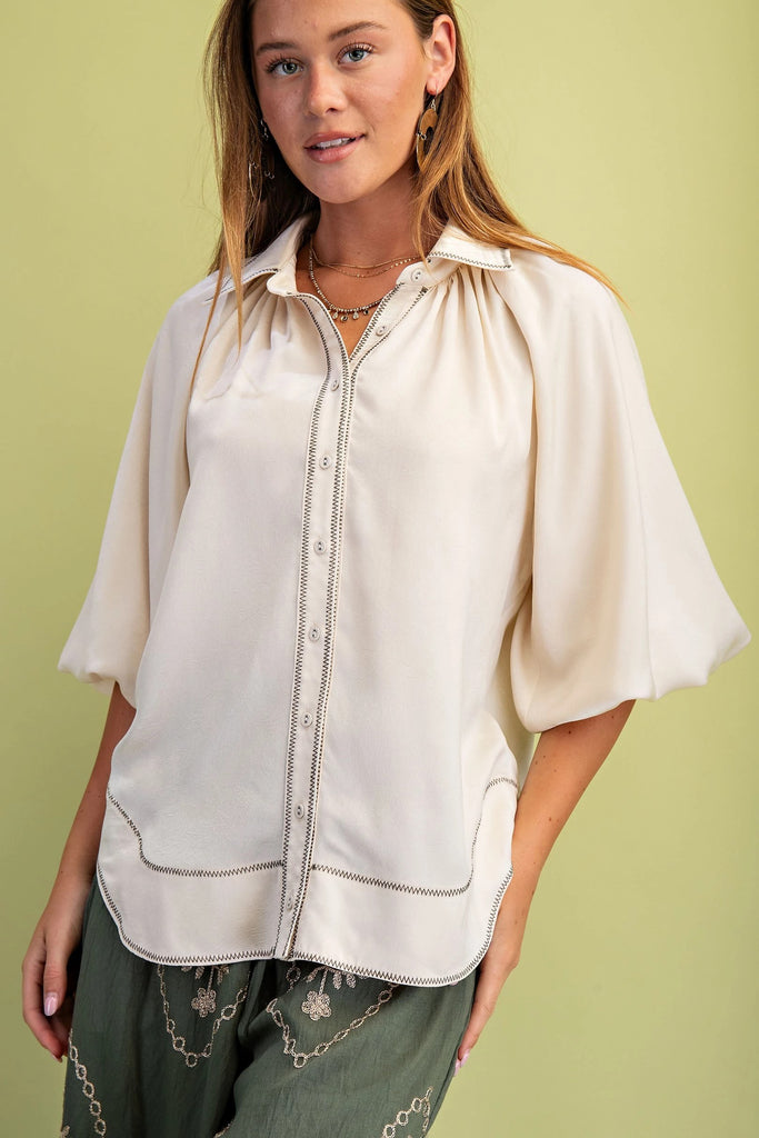 A cream satin button-down blouse with three-quarter balloon sleeves and black zigzag contrast stitching on the front placket and hem.