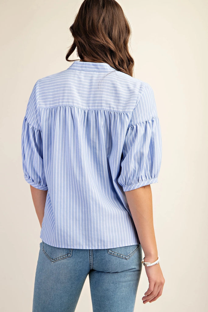 A back shot of a brunette woman wearing a blue and white striped blouse with a split v-neck and quarter-length bubble sleeves. She is also wearing blue jeans and simple pearl jewelry. The background is a solid light beige.