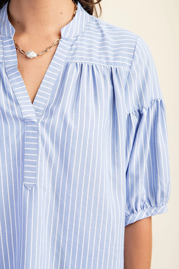 A product shot of a woman wearing a blue and white striped blouse with a split v-neck and quarter-length bubble sleeves.  The background is a solid light beige.