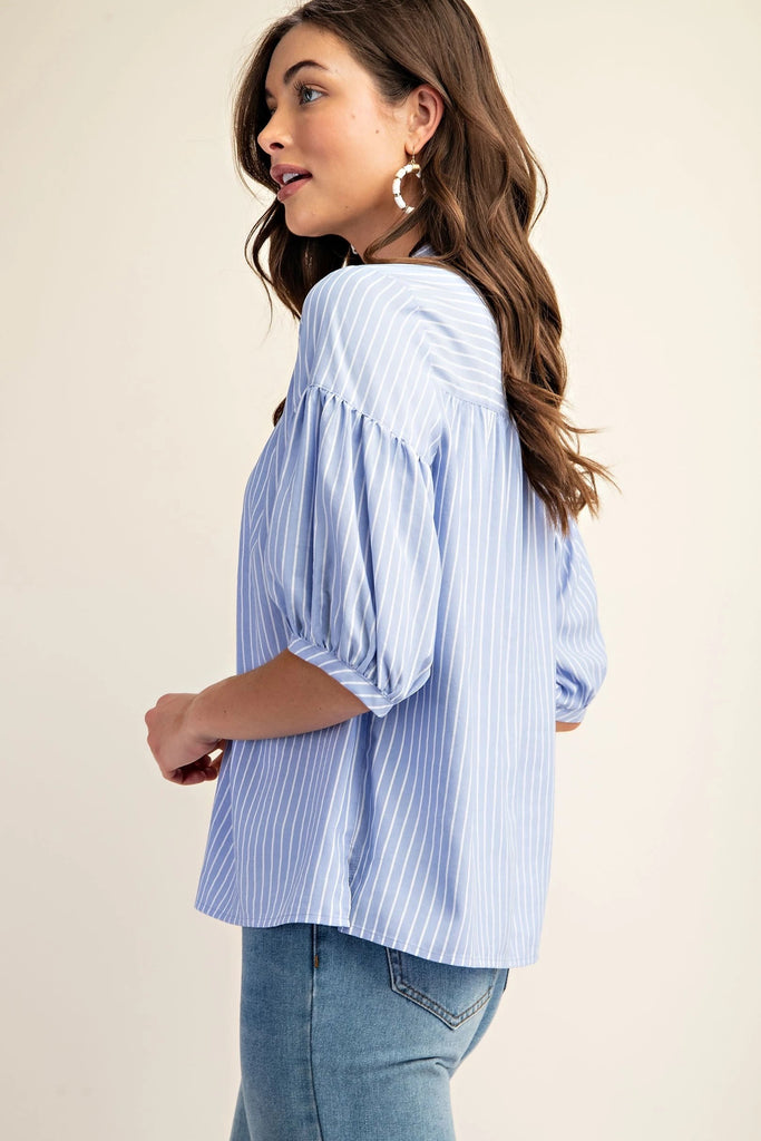 A product shot of a  brunette woman wearing a blue and white striped blouse with a split v-neck and quarter-length bubble sleeves. She is also wearing blue jeans and simple pearl jewelry. The background is a solid light beige.