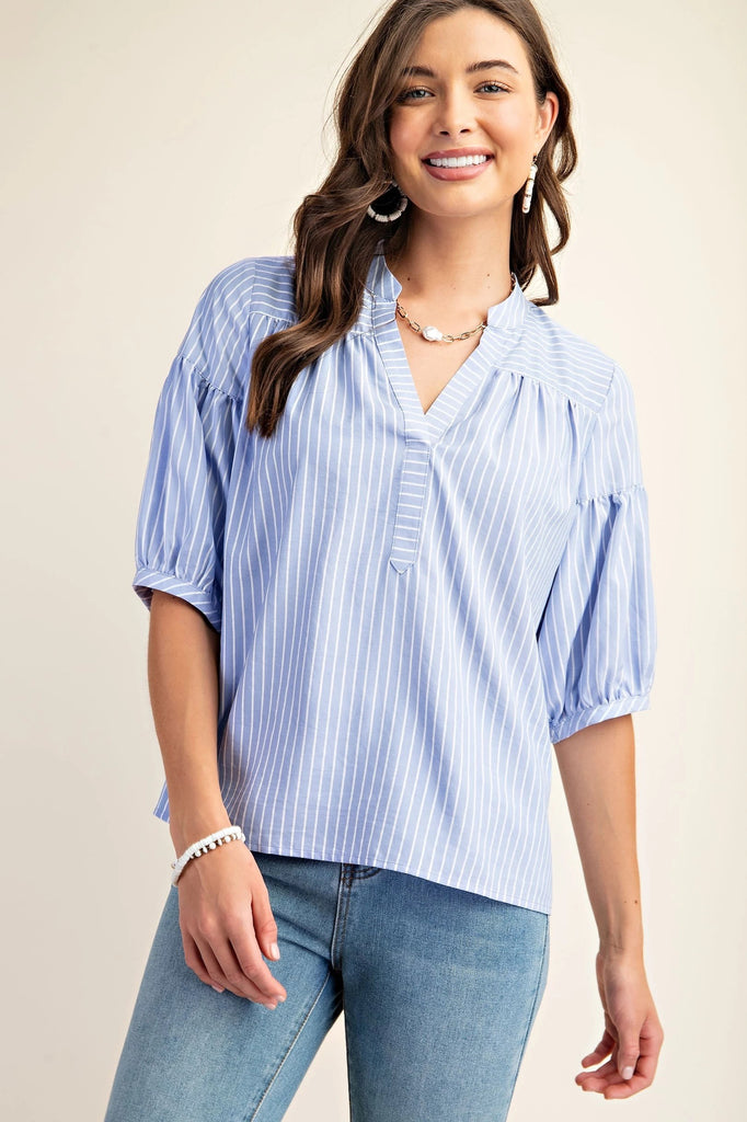 A product shot of a smiling brunette woman wearing a blue and white striped blouse with a split v-neck and quarter-length bubble sleeves. She is also wearing blue jeans and simple pearl jewelry. The background is a solid light beige.