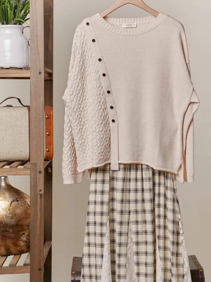 A cream-colored, long-sleeve sweater with a boat neck and decorative buttons down the side is shown hanging, styled over a plaid midi skirt. The sweater features a textured knit panel on one side.