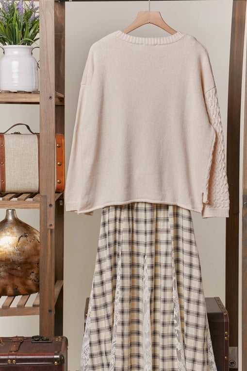 A cream-colored, long-sleeve sweater with a boat neck and decorative buttons down the side is shown hanging, styled over a plaid midi skirt. The sweater features a textured knit panel on one side.