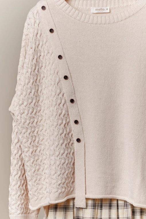 A cream-colored, long-sleeve sweater with a boat neck and decorative buttons down the side is shown hanging, styled over a plaid midi skirt. The sweater features a textured knit panel on one side.