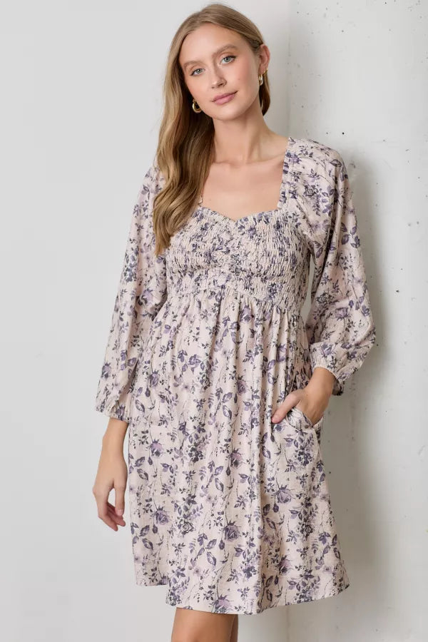 A natural-colored mini dress with a dark floral print. It has a square neckline, a smocked bodice, and long balloon sleeves with elastic cuffs.