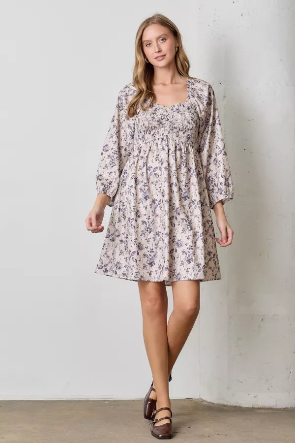 A natural-colored mini dress with a dark floral print. It has a square neckline, a smocked bodice, and long balloon sleeves with elastic cuffs.