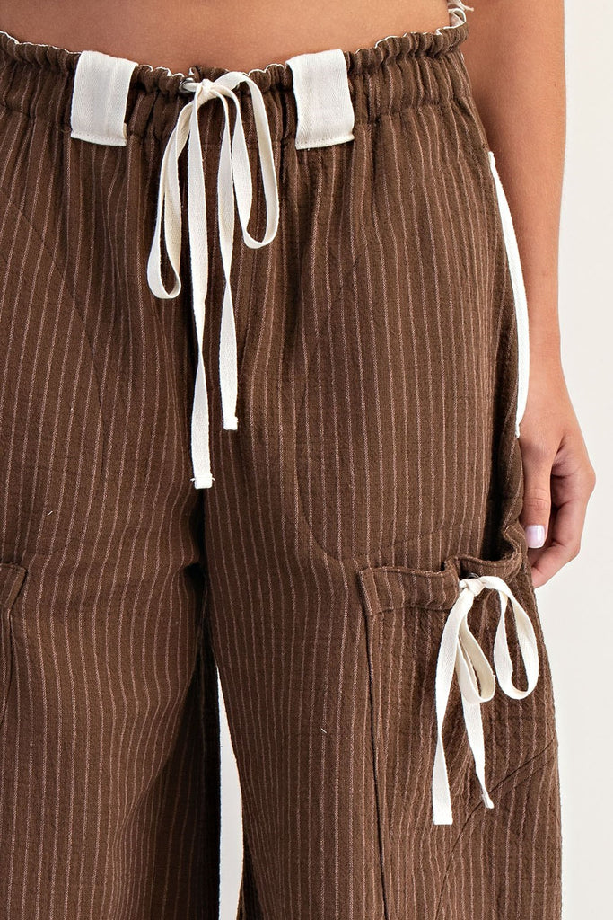 Model wearing relaxed-fit brown striped jogger pants with an elastic waistband and contrasting white drawstring ties. Pants feature utility-style flap pockets, front and back patch pockets, and gathered elastic hems, styled with open-toe logo slides.
