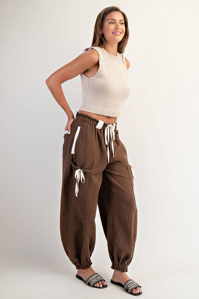 Model wearing relaxed-fit brown striped jogger pants with an elastic waistband and contrasting white drawstring ties. Pants feature utility-style flap pockets, front and back patch pockets, and gathered elastic hems, styled with open-toe logo slides.