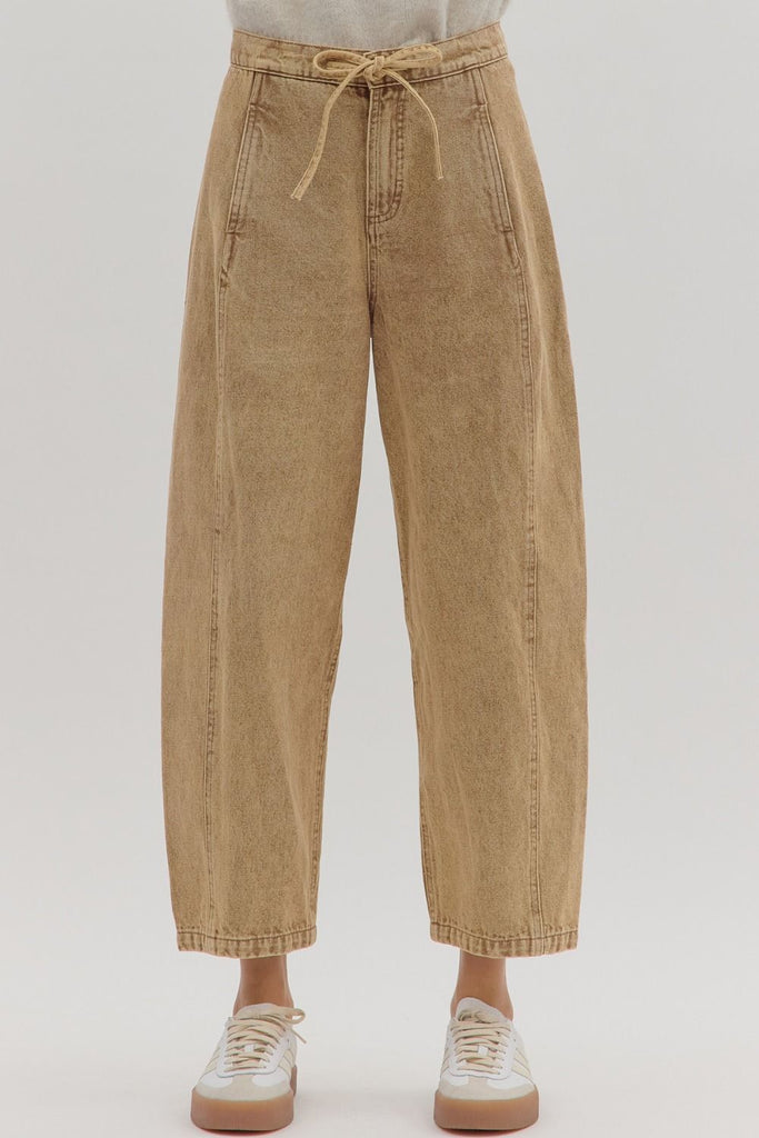 A person models a pair of high-waisted, tan-colored denim pants with a drawstring waist, button closure, and vertical seam details down the front of each leg.