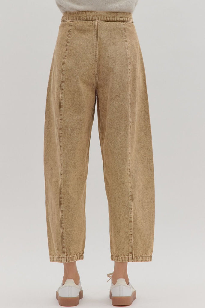 A person models a pair of high-waisted, tan-colored denim pants with a drawstring waist, button closure, and vertical seam details down the front of each leg.