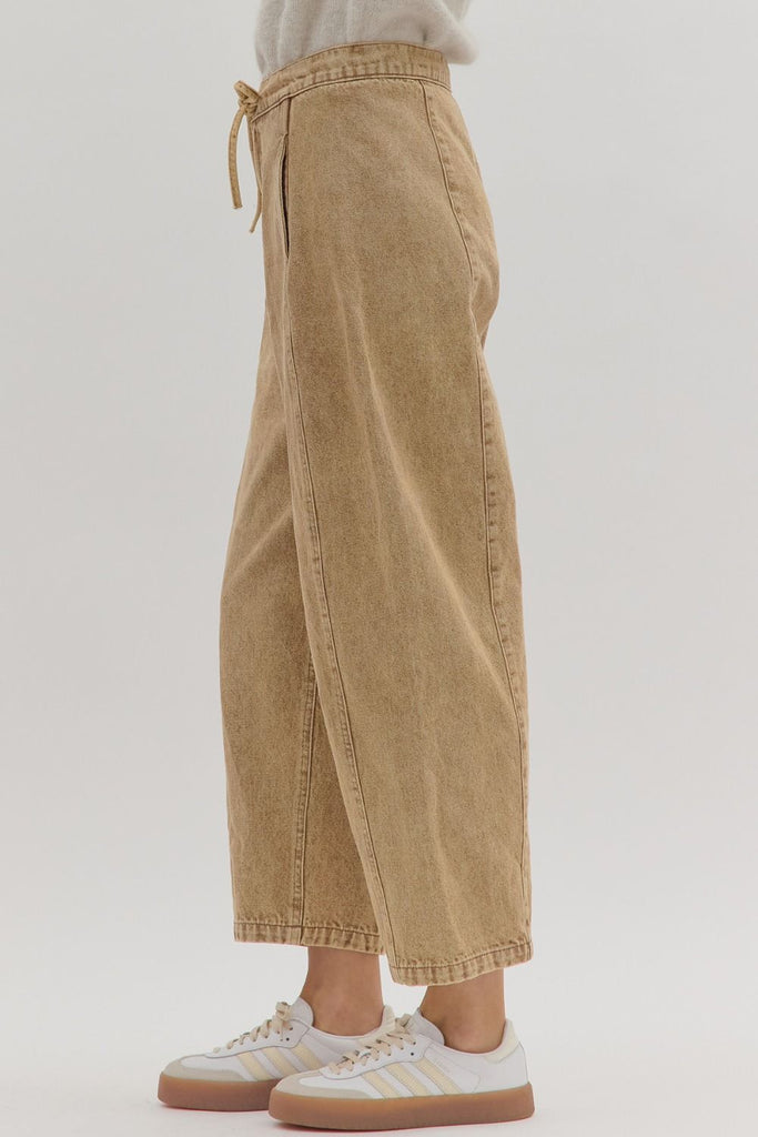 A person models a pair of high-waisted, tan-colored denim pants with a drawstring waist, button closure, and vertical seam details down the front of each leg.