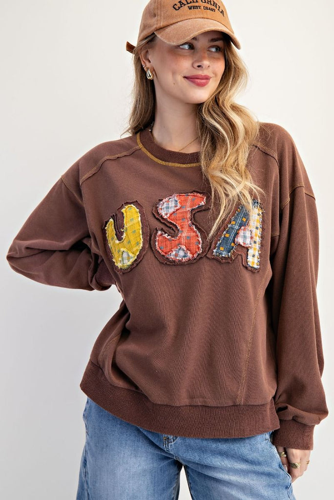 A mocha-colored washed terry pullover with a relaxed fit, featuring patchwork "USA" lettering on the front, dropped shoulders, ribbed cuffs and hem, and contrast topstitching at the neckline.