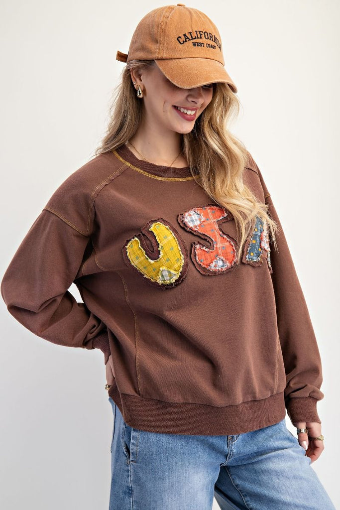A mocha-colored washed terry pullover with a relaxed fit, featuring patchwork "USA" lettering on the front, dropped shoulders, ribbed cuffs and hem, and contrast topstitching at the neckline.