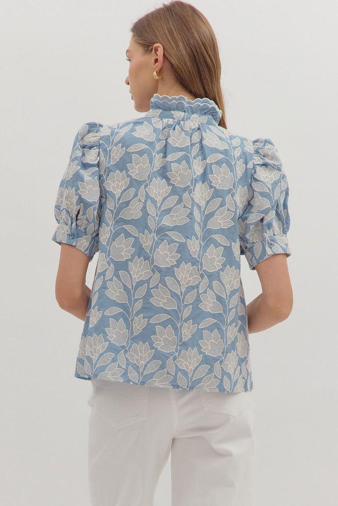 A chambray-colored short puff-sleeve top with white botanical embroidery, a ruffled stand-up collar, and a button-down front.