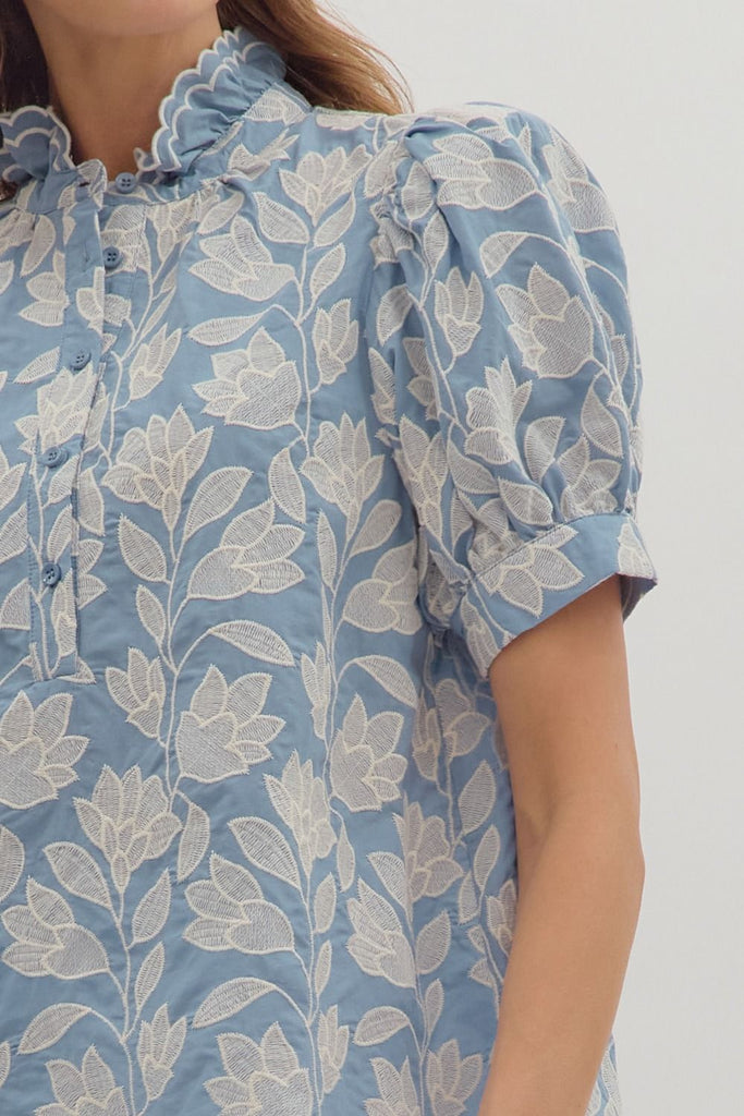 A chambray-colored short puff-sleeve top with white botanical embroidery, a ruffled stand-up collar, and a button-down front.