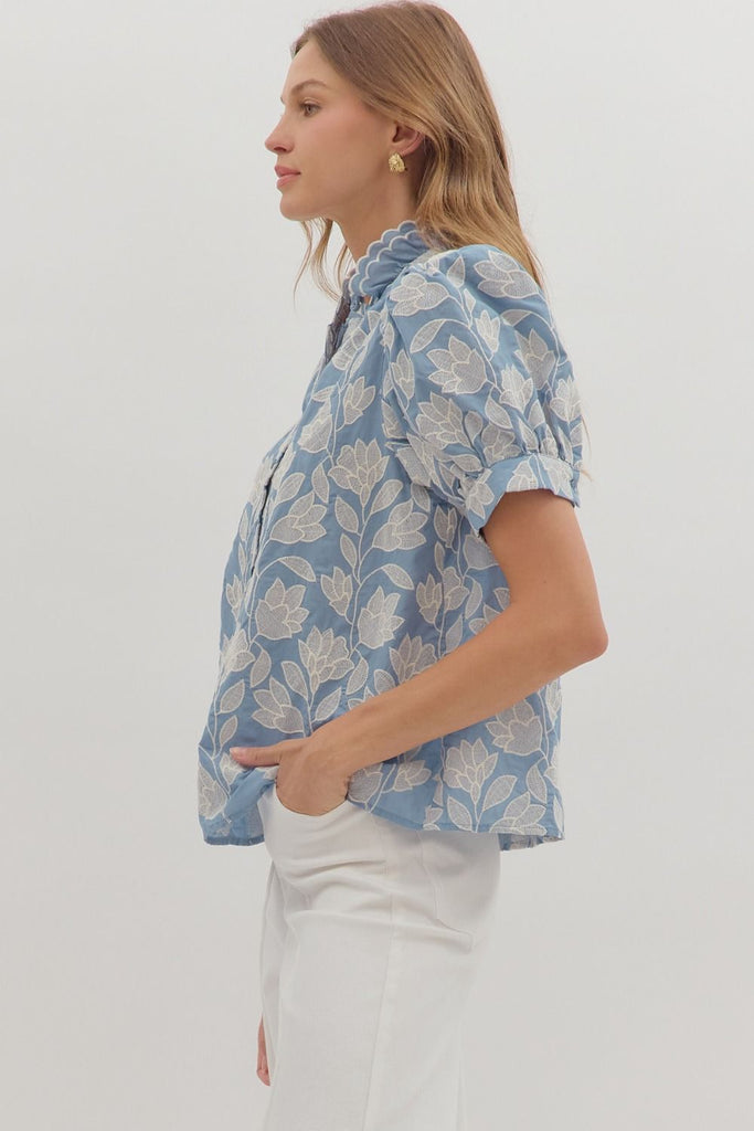 A chambray-colored short puff-sleeve top with white botanical embroidery, a ruffled stand-up collar, and a button-down front.