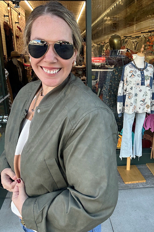 Woman wearing sunglasses and a green jacket in a store setting with mannequins and clothing.